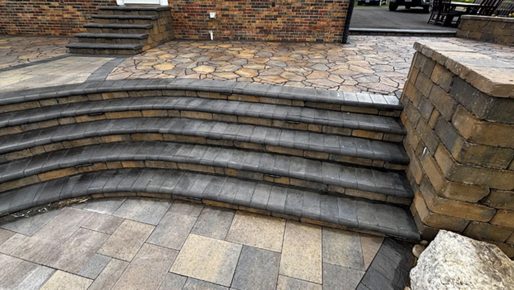 Wide curved outdoor steps constructed with gray and tan paver stones, connecting different levels of patio area with decorative flagstone pattern, professional hardscaping design