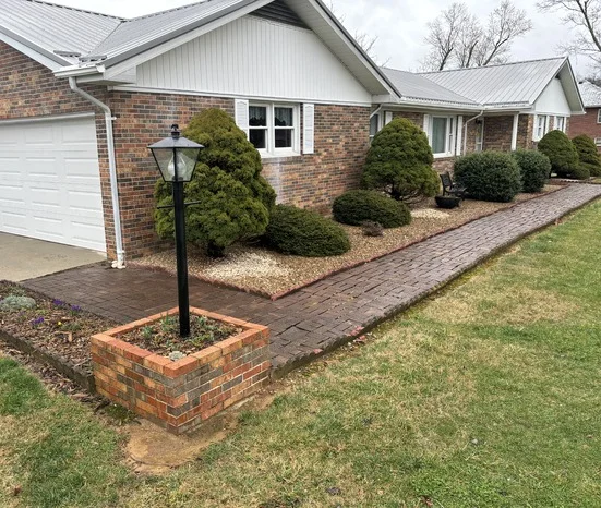 Professional brick paver walkway with matching brick lamp post planter box, manicured evergreen shrubs in mulched beds, complementing brick home with white trim and metal roof