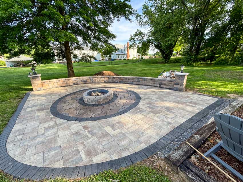 Paver patio with a curved seating wall and a stone fire pit on open ground, mature trees and lawn beyond