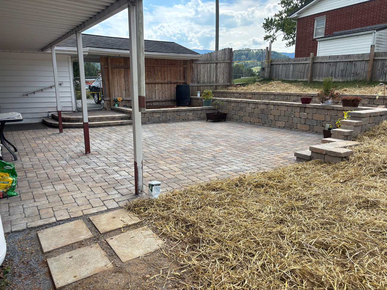 Paver patio beside a stacked block retaining wall holding a planted slope, with steps leading up to the higher level