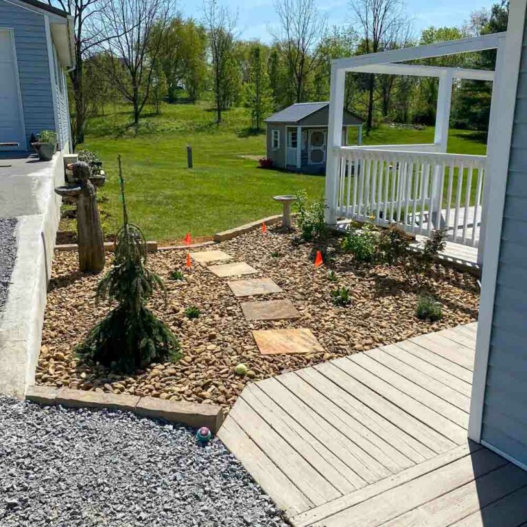 A view from a house's front door showcasing a landscaped backyard with a variety of rocks and plants creating a rock garden. A wooden deck extends from the house, leading towards the rock garden and providing a space for relaxation outdoors.